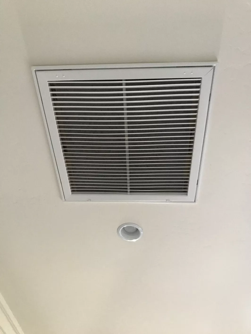 An air vent located next to a can light on an all white ceiling.