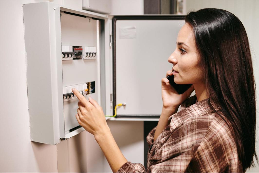A young brunette woman wearing a brown flannel inspecting her electrical panel’s breaker box.
