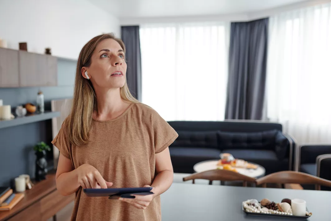 A dirty blonde woman wearing a tan shirt, with an air pod in her right ear, looking up, and holding a smart device tablet in her kitchen.