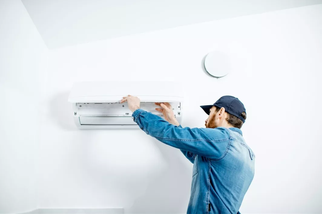 A man wearing a jean shirt and blue baseball cap, inspecting an ac unit affixed to a white wall.