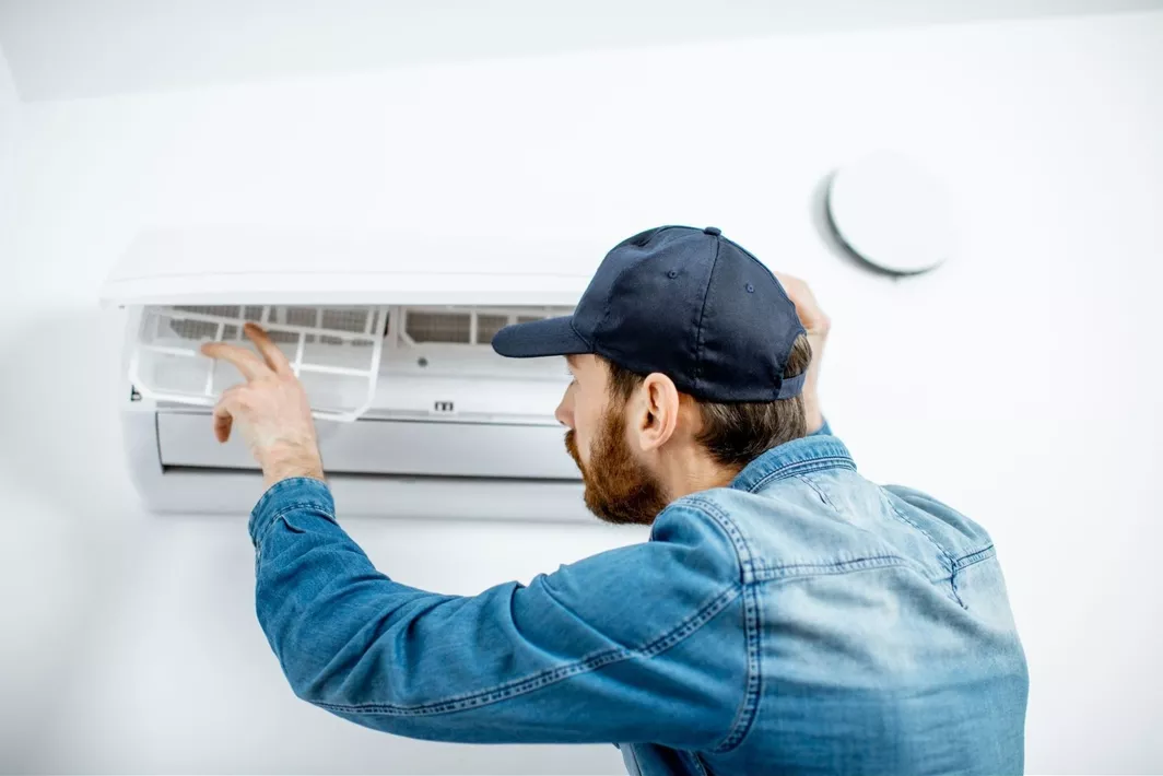 A man wearing a jean long sleeve shirt and blue baseball cap changing out an air filter in a wall AC unit.