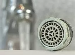 Closeup of a shower head covered in hard water stains with a faucet with running water in the background