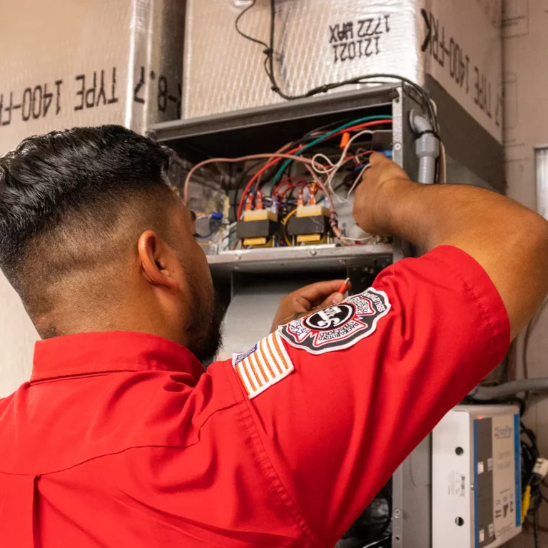 Technician in red uniform inspecting internal components of a heating system with visible wiring and circuit board.